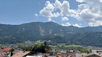 Panoramablick über die Dächer einer Stadt mit majestätischen Bergen im Hintergrund unter blauem Himmel.