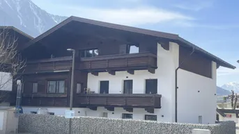Traditionelles Gebäude mit Holzbalkonen und weißer Fassade vor alpiner Bergkulisse.