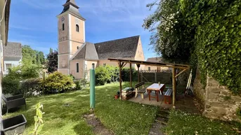 Grüner Garten mit überdachtem Sitzbereich und Blick auf die Kirche.