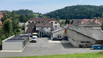 Außenansicht des Anwesens mit mehreren Gebäuden, großem Parkplatz und grüner Hügellandschaft im Hintergrund.