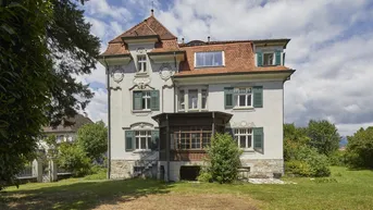 Historische Villa mit markanter Architektur, Walmdach und gepflegtem Vorgarten unter blauem Himmel.