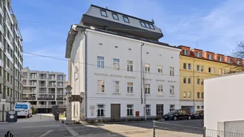 Weißes Mehrfamilienhaus mit modernem Dachausbau und umliegenden Gebäuden unter blauem Himmel.