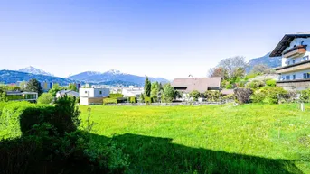Weitläufiger Blick über grüne Wiesen und Häuser mit schneebedeckten Bergen im Hintergrund.