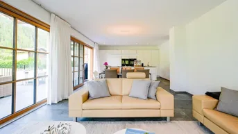 Helles, offenes Wohnzimmer mit beigen Ledersofas, Blick auf die Terrasse und moderne Küche im Hintergrund.