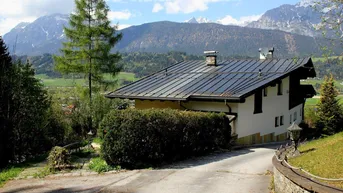 Haus mit Photovoltaikanlage auf dem Dach und Zufahrt, umgeben von grüner Natur und Bergen.