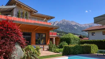 Modernes Haus mit Pool und Garten, umgeben von einer beeindruckenden Berglandschaft unter blauem Himmel.