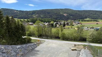 Ländliche Umgebung mit Häusern, grünen Feldern und bewaldeten Bergen im Hintergrund.