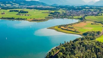 Luftaufnahme einer malerischen Seenlandschaft mit Bergen im Hintergrund und grünen Feldern.