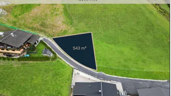 Luftaufnahme eines Baugrundstücks von 543 m² Größe, eingebettet in eine grüne Hügellandschaft mit umliegenden Häusern.