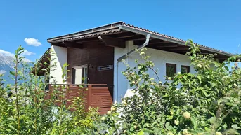 Hausfassade mit Balkon und üppiger grüner Bepflanzung vor einer Bergkulisse.