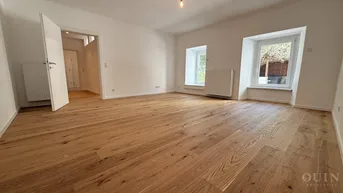 Helles Wohnzimmer mit neuem Holzparkettboden und zwei Fenstern, die viel Tageslicht hereinlassen.