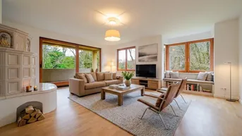 Helles Wohnzimmer mit großem Kachelofen, Holzboden und Zugang zu einem Balkon mit Grünblick.