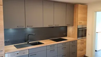 Moderne Küche mit grauen Fronten, Holzarbeitsplatte, integrierten Geräten und dunklem Fliesenboden.