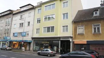 Ansicht eines gepflegten Mehrfamilienhauses mit hellgrüner Fassade und Geschäften im Erdgeschoss in einer städtischen Umgebung.