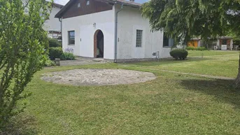 Weißes Einfamilienhaus mit großem, gepflegtem Garten und altem Baumbestand unter bewölktem Himmel.
