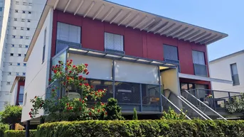 Modernes Gebäude mit markanter roter Fassade, verglaster Loggia und gepflegter Hecke im Vordergrund unter blauem Himmel.