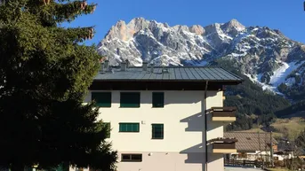 Seitenansicht des Gebäudes mit Balkonen und einem beeindruckenden Bergpanorama unter blauem Himmel.