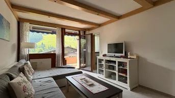 Gemütliches Wohnzimmer mit Holzbalkendecke, Teppichboden und Zugang zum Balkon mit Bergblick.