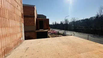 Rohbau eines Gebäudes mit unverputzten Ziegelwänden und einer angrenzenden Baustelle unter blauem Himmel.