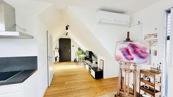 Helles Wohnzimmer mit Dachschrägen, Holzboden und integrierter Küchenzeile.