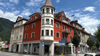Historisches Gebäude mit markantem Turm und roter Fassade an einer belebten Straßenecke mit Bergblick.