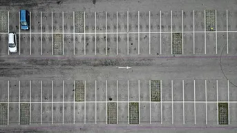 Luftaufnahme eines Parkplatzes mit markierten Stellplätzen, zwei Autos und begrünten Zwischenräumen.