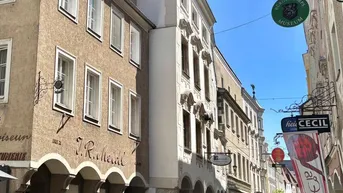 Historische Gebäude mit Geschäften im Erdgeschoss und gepflasterter Straße unter blauem Himmel in einer Altstadt.