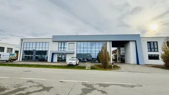 Modernes Geschäftsgebäude mit großer Glasfassade und Parkplätzen davor unter bewölktem Himmel.
