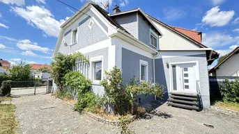 Graues Einfamilienhaus mit weiß abgesetzten Fensterrahmen und einer modernen Eingangstür unter blauem Himmel.