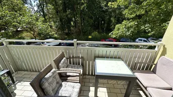 Möblierter Balkon mit Blick ins Grüne und auf parkende Autos unter strahlendem Sonnenschein.