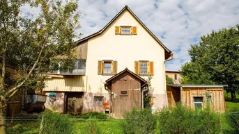 Traditionelles Landhaus mit beiger Fassade, Holzfensterläden und Satteldach, umgeben von viel Grün.