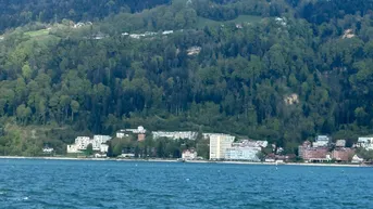 Panoramablick über den See mit Uferbebauung und bewaldeten Hügeln im Hintergrund.