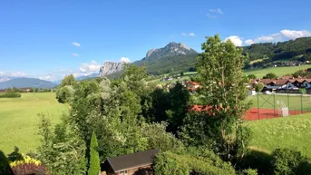 Weitläufige Landschaft mit grünen Feldern, Bäumen und Bergen unter blauem Himmel.