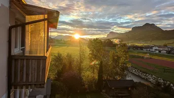 Sonnenuntergang über einer ländlichen Landschaft mit Bergen und einem Balkon im Vordergrund.