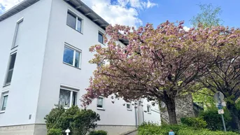 Modernes Mehrfamilienhaus mit weißer Fassade und blühendem Baum im Vorgarten unter blauem Himmel.