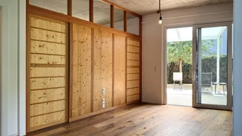 Heller Wohnraum mit Holzboden, Sichtbetondecke und großem Fenster zur Terrasse.