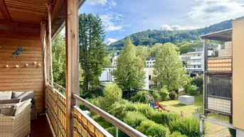Geräumiger Balkon mit Holzverkleidung, Sitzgelegenheiten und Blick ins Grüne mit Spielplatz.