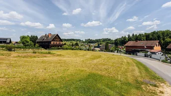 Ländliche Umgebung mit einem unbebauten Grundstück und Wohnhäusern im Hintergrund unter blauem Himmel.