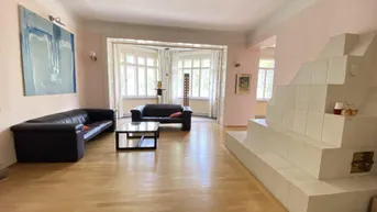 Geräumiges Wohnzimmer mit hellem Parkettboden, einem traditionellen Kachelofen und großen Erkerfenstern.