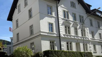 Weißes Mehrfamilienhaus mit klassischer Architektur, umgeben von einer gepflegten Hecke und parkenden Autos.