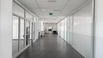Langer, heller Büroflur mit Glaswänden und dunklem Fliesenboden, der zu verschiedenen Büros führt.