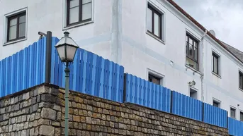Weißes Gebäude mit dunklen Fenstern und einem markanten blauen Zaun auf einer Steinmauer.