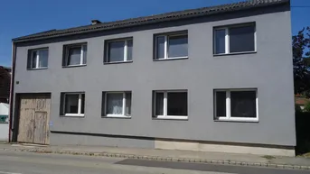 Ansicht der grauen Fassade eines zweistöckigen Gebäudes mit mehreren Fenstern und einer großen Holztür.