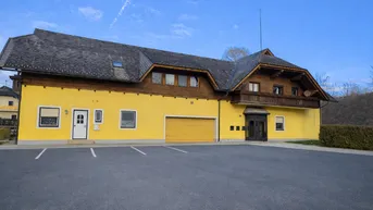 Gelbes Gebäude mit Holzverkleidung, Satteldach und großem Parkplatz davor unter blauem Himmel.