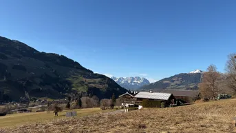 Weitläufige Landschaft mit Bergen im Hintergrund und einem traditionellen Gebäude im Tal unter blauem Himmel.