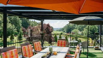 Überdachte Terrasse mit Esstisch und Stühlen, die einen weiten Blick über die grüne Landschaft bietet.