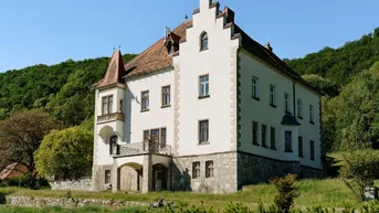 Historisches Gebäude mit weißer Fassade und rotem Ziegeldach, umgeben von grüner Natur.