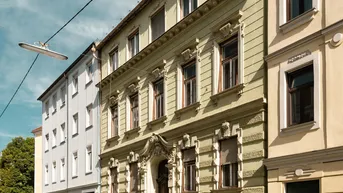 Historisches Gebäude mit verzierter Fassade und Rollläden in einer städtischen Umgebung.