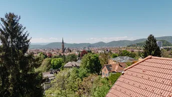 Panoramablick über die Stadt mit Kirchturm, grünen Hügeln und Bergen im Hintergrund.