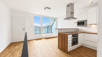 Heller offener Wohnbereich mit moderner Einbauküche, Holzboden und Zugang zu einem kleinen Balkon.
